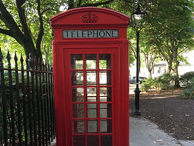 A bright red phone booth in London awaits its next occupant next to a tall iron fence and a healthy tree. 