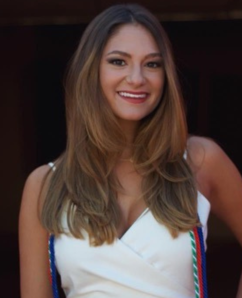 Laura wears a white dress with graduation tassels. She stands against a dark background smiling with long brown hair.