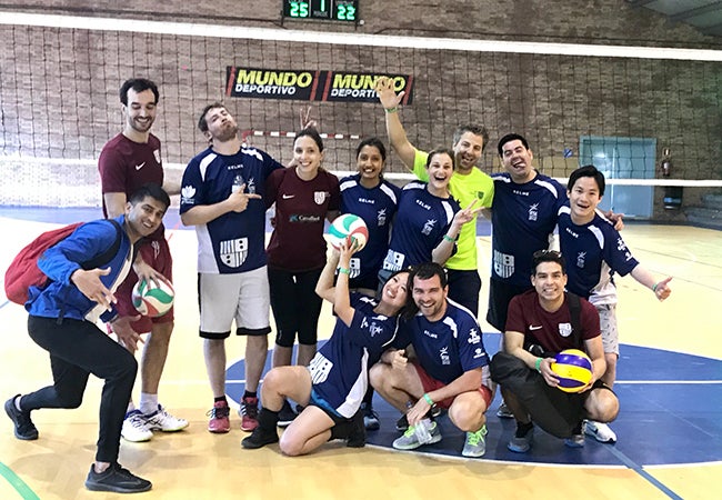 Business school students dressed in uniforms pose with a volleyball in front of a net. 