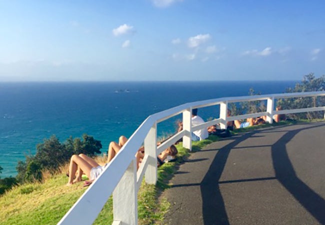 Students look out at the sea while laying in the grass on a hillside next to a road with a while guardrail to catch some rays during a break from studies. 