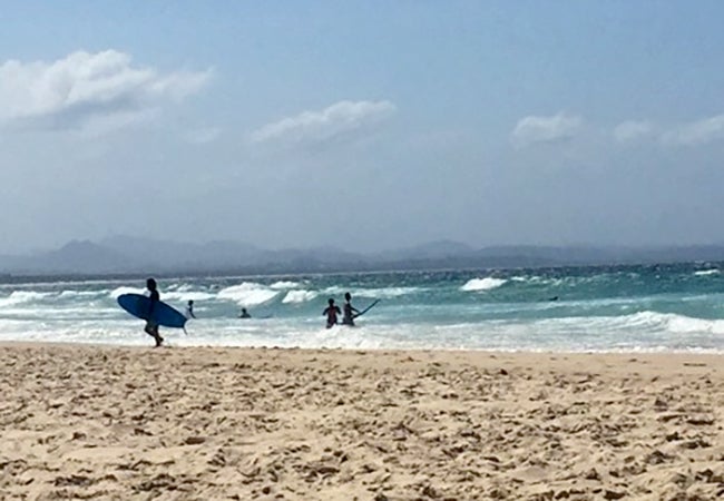 Surfers carry their boards out to the beach as waves crest nearby on a sunny day in Australia. 