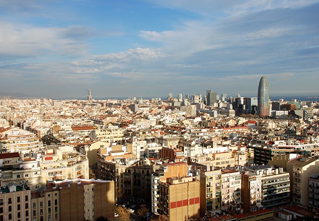 A scenic view of Barcelona shows many low-rise tan buildings under red rooftops on a blue-sky day.