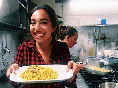 Undergraduate business student Alexandra Grieco holds a plate of pasta while standing in an industrial kitchen as steam rises in the background.