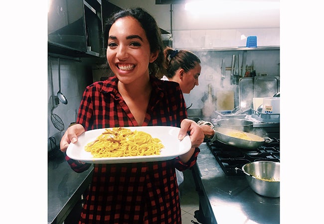 Undergraduate business student Alexandra Grieco holds a plate of pasta while standing in an industrial kitchen as steam rises in the background.