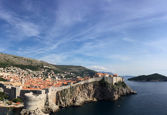 An aerial view shows a stone wall surrounding the city of Dubrovnik, perched on a cliff by the sea. 