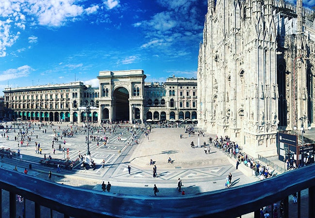 Pedestrians mill about a large city square in Milan surrounded by magnificent stone buildings. 