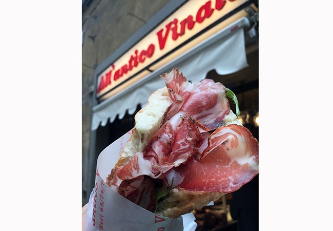 Someone holds up a panini purchased from All’antico Vinaio in front of the restaurant's sign. 