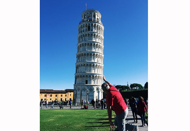 A man in a red shirt holds up a hand as if to correct the posture of the Leaning Tower of Pisa.