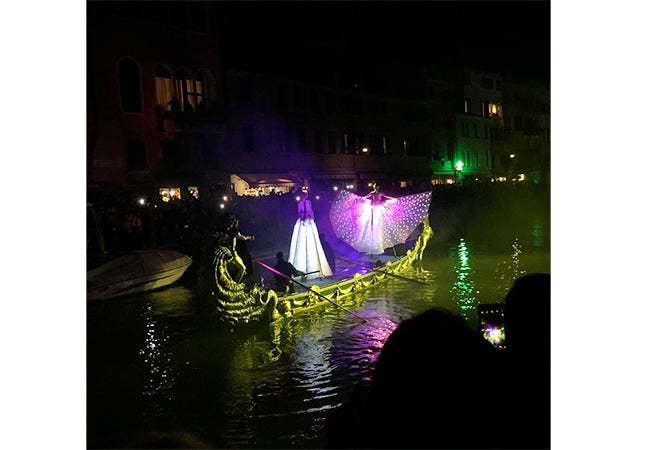 Two figures in capes and dresses appear to float atop a gondola, surrounded by a purple glow, during Carnival in Venice. 