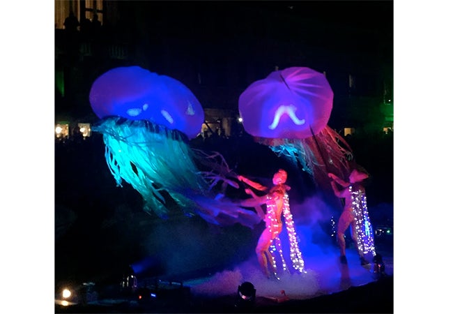 Two figures shake large objects that resemble jellyfish, illuminated in blue and green light, during Carnival in Venice, Italy.