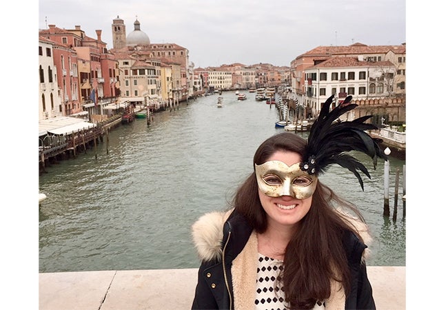 MBA student Ashley Grand wears a shiny gold mask while standing in front of a waterway lined with buildings in Italy.
