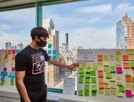 Student participating in the Design Sprint pointing to sticky notes with Brooklyn Bridge outside the window