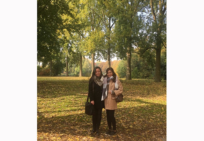 MBA student Kimberly Rodriguez and a friend pose beneath a stand of large trees in a park during the fall. 
