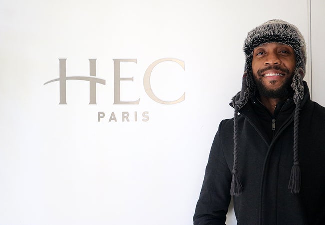 MBA student Calvin Mack wears a warm hat and stands in front of a sign that says "HEC Paris." 