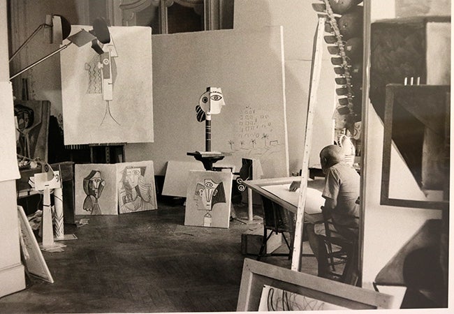 A black-and-white photo shows Picasso hunched over a painting in his studio while surrounded by works in progress. 