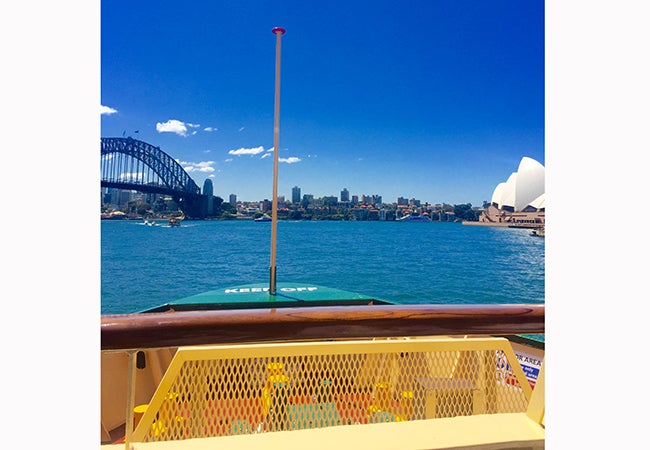 The bow of a boat rises before the blue sea and sky with a cityscape in the distance. 