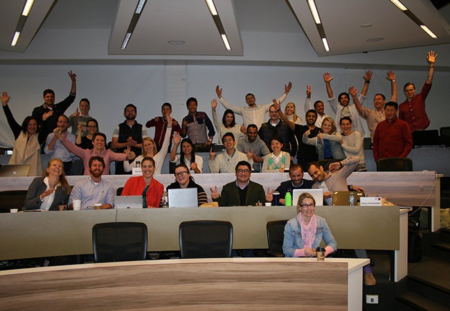 A large group of students hold up their hands while seated and standing throughout a classroom at Melbourne Business School. 