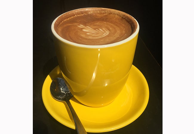 A flat white served in a bright yellow mug with a leaf pattern on top. 