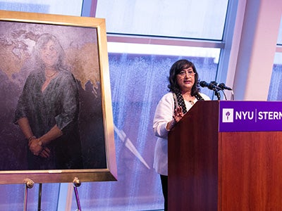 Dean Menon speaks at podium with portrait of her displayed on her right