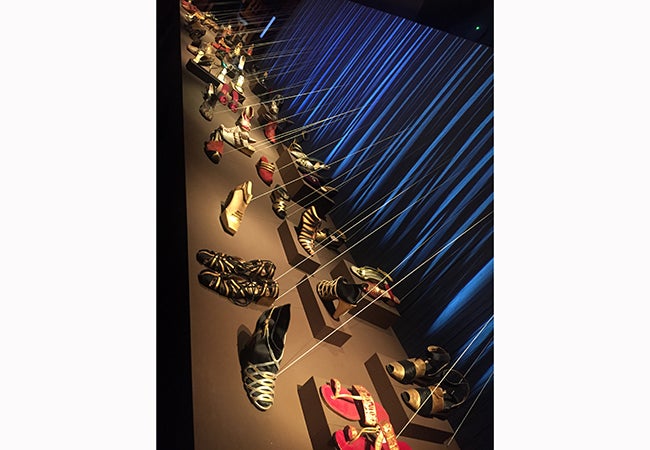 Pairs of colorful sandles, strappy heels, and flats are shown on display at a museum exhibit. 