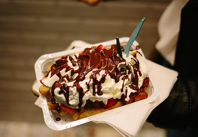 Someone holds out a container with a Belgian waffle topped with generous helpings of fruit and whipped cream and drizzled with chocolate. 