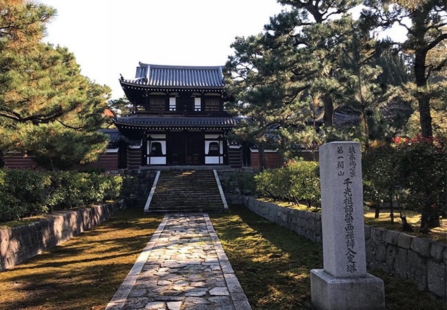 A dark brown building in Japan is surrounded by trees and behind a sign with Japanese characters. 