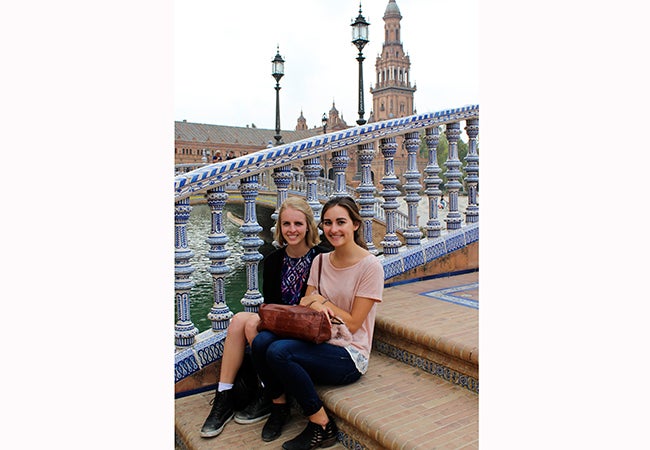 Undergraduate student Michelle Enkerlin and a friend sit on a stoop with an ornately-decorated railing behind them. 