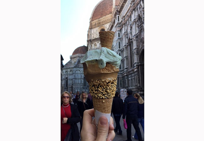 Someone holds a cone of gelato in front of a stone city building. 