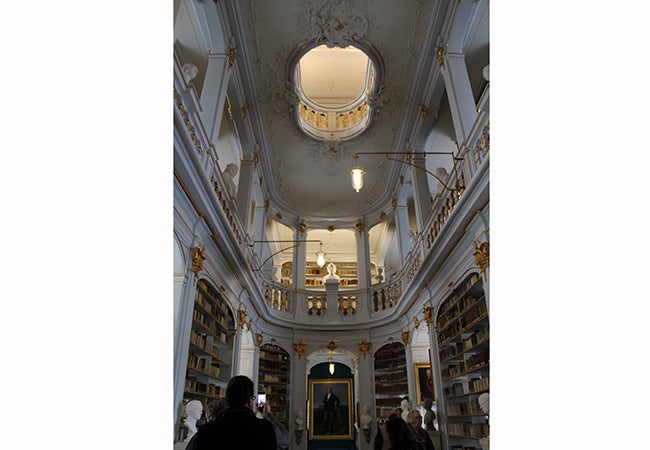 A large portrait hangs on the first level of a three-story library decorated with gold fixtures. 