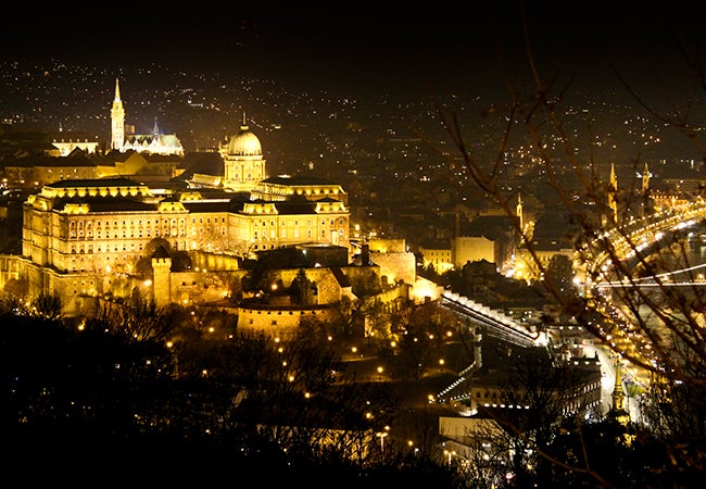 An aerial view of Budapest shows several large stone buildings and complexes lit up a night. 