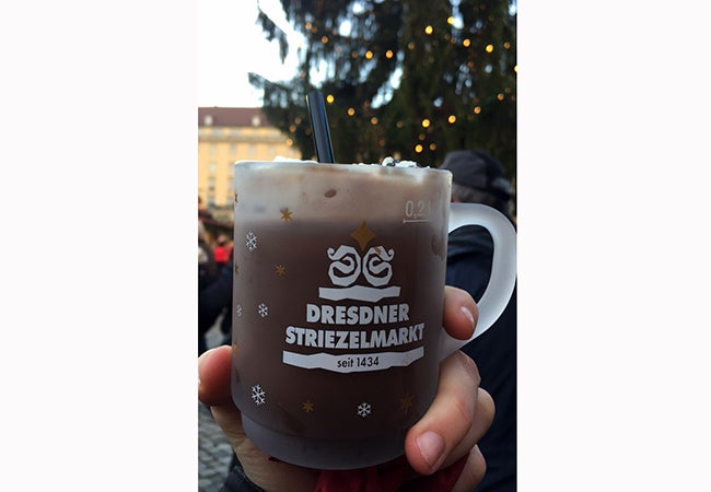 A hand holds up a glass mug of hot chocolate with a black straw. 