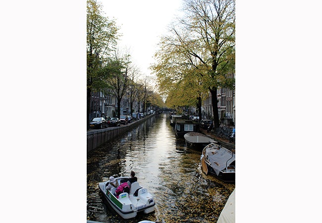 Three passengers begin to steer a paddle boat down a canal in Amsterdam covered in leaves from nearby trees. 