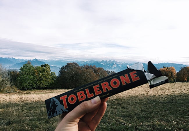 A woman's hand holds up a Toblerone candy bar in front of a mountainous scene. 