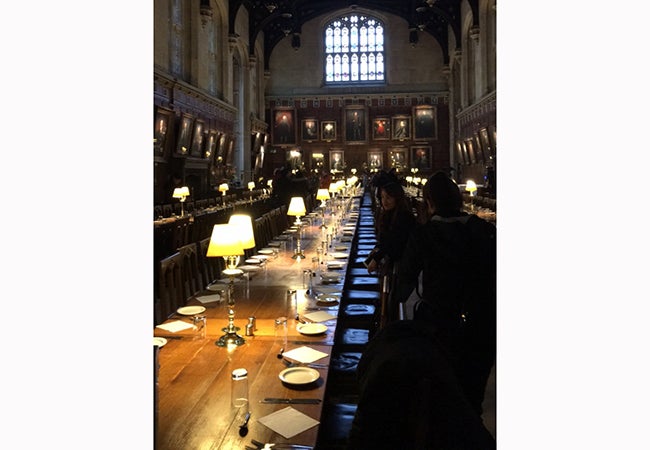 A large dining hall at Oxford includes portraits of university leaders and desk lamps illuminating long wooden tables. 