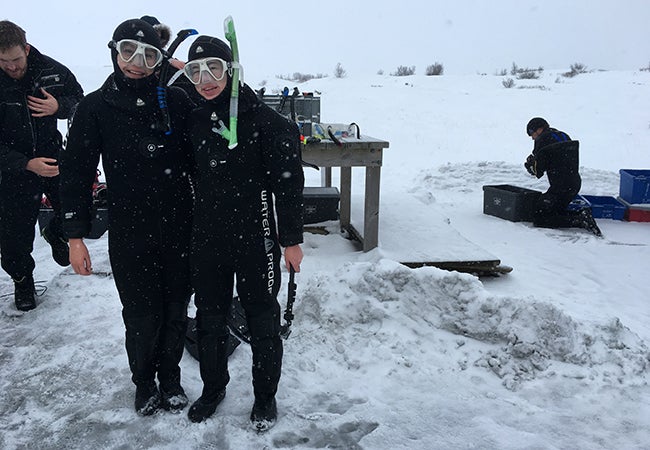 Two business students dressed in black wetsuits and snorkeling gear pose in front of an icy opening in the water on a frigid day.