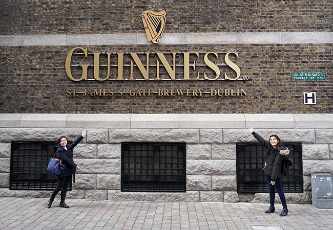 Undergraduate business student Natasha Lim and her friend point to a large golden sign that says "Guiness" on the brick exterior of a brewery.. 