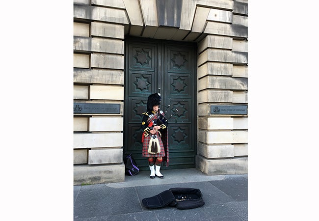 A bagpipe player wearing traditional dress stands in a doorway to play to passersby behind a suitcase open to collect coins.