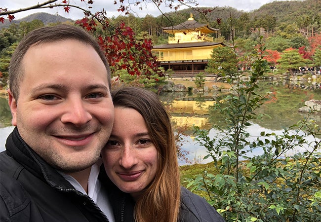 Part-time MBA student Ryan and his girlfriend stand in a garden in Japan with a vivid red tree and a golden structure in the background. 