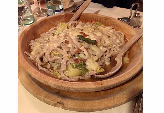 A close-up of a dish called pizzoccheri served in a wooden bowl with a wooden spoon. 