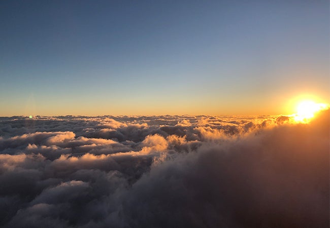 A scenic photo shows a calm blue sky above the clouds as the sun sets or rises in the distance. 