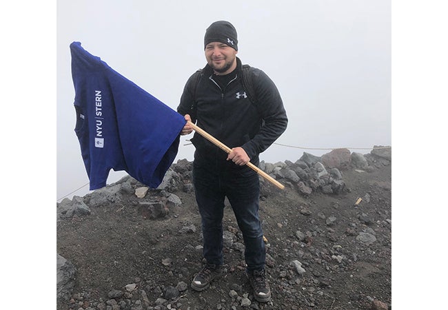 Ryan McClaskey holds a stick with an NYU t-shirt draped over the end like a flag while hiking on a chilly day.