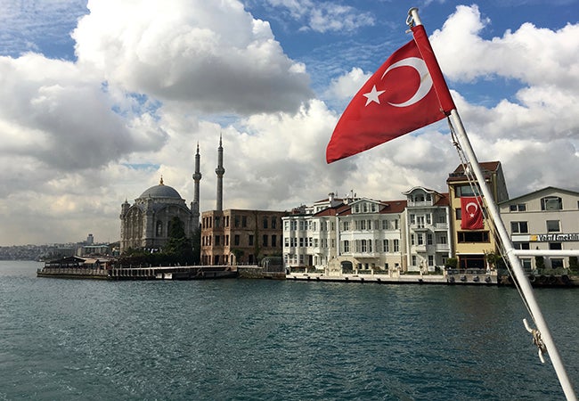 The Turkish flag flies before a body of water and several city buildings in Istanbul.