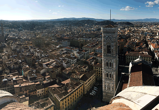 A tall tower sticks out among the other buildings in an aerial photo of the city of Florence, Italy on a clear day. 