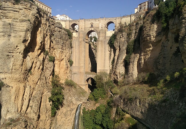 An ancient aqueduct built into a stone hillside towers above a small stream on a clear day. 