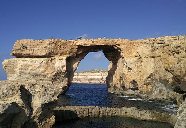 Visitors cross over a large stone arch surrounded on three sides by cliffs and water on a bright, sunny day in Malta. 