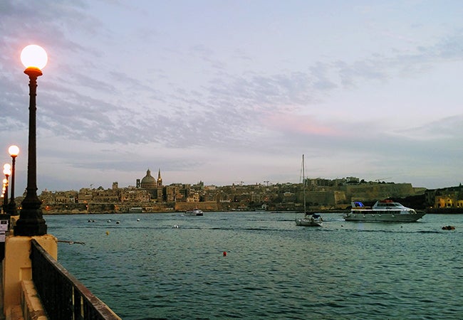 A seaside view of Malta shows small boats before a low-slung city of stone buildings in the distance.