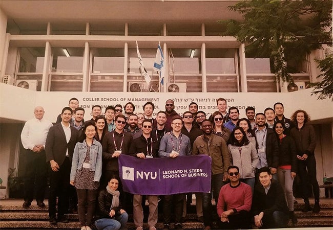 A group of MBA students hold a purple banner emblazoned with Stern's logo while standing on the stairs outside of a classroom building.