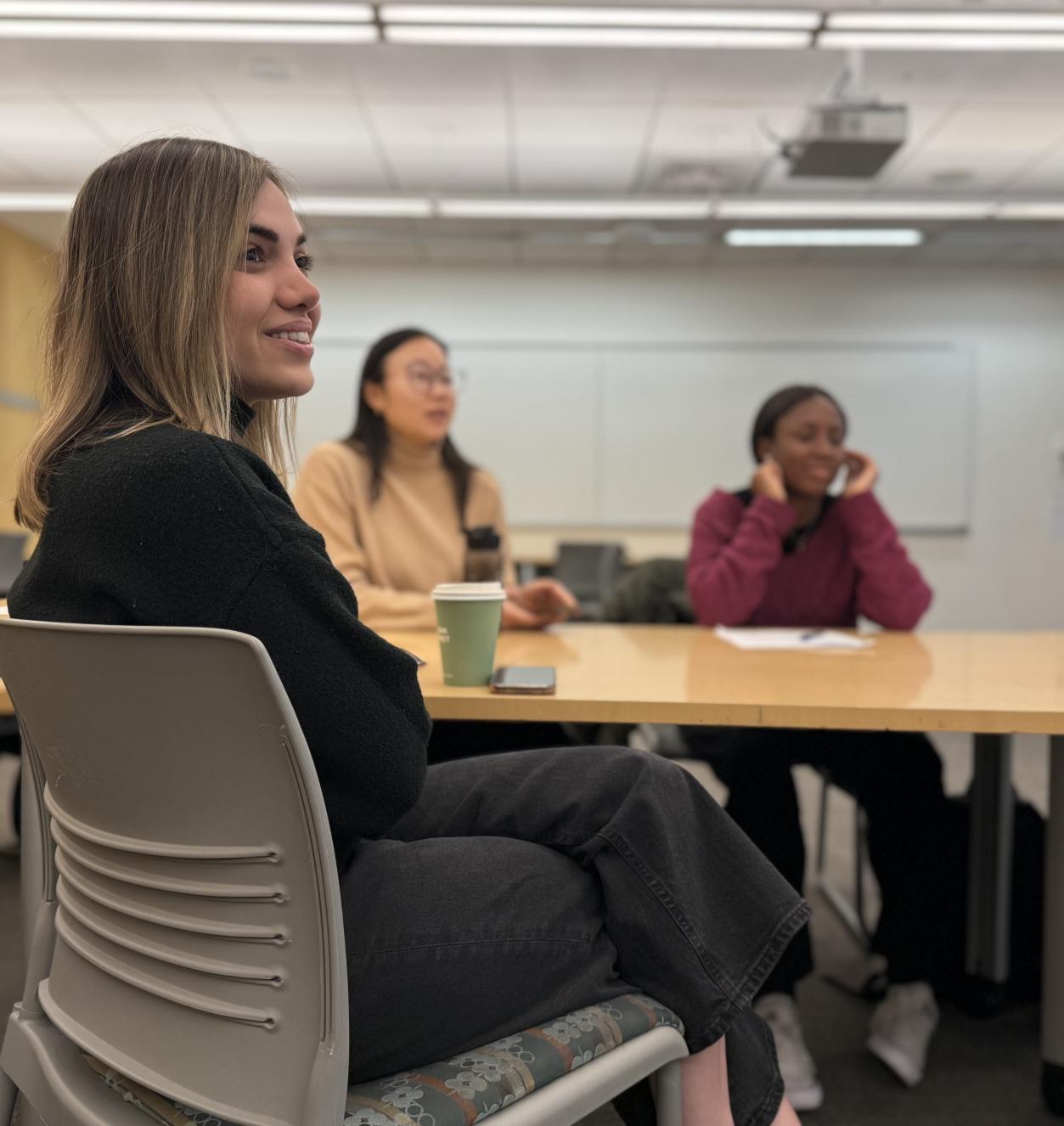 Three feminine-presenting students sit in a half-moon and smile in a classrom.
