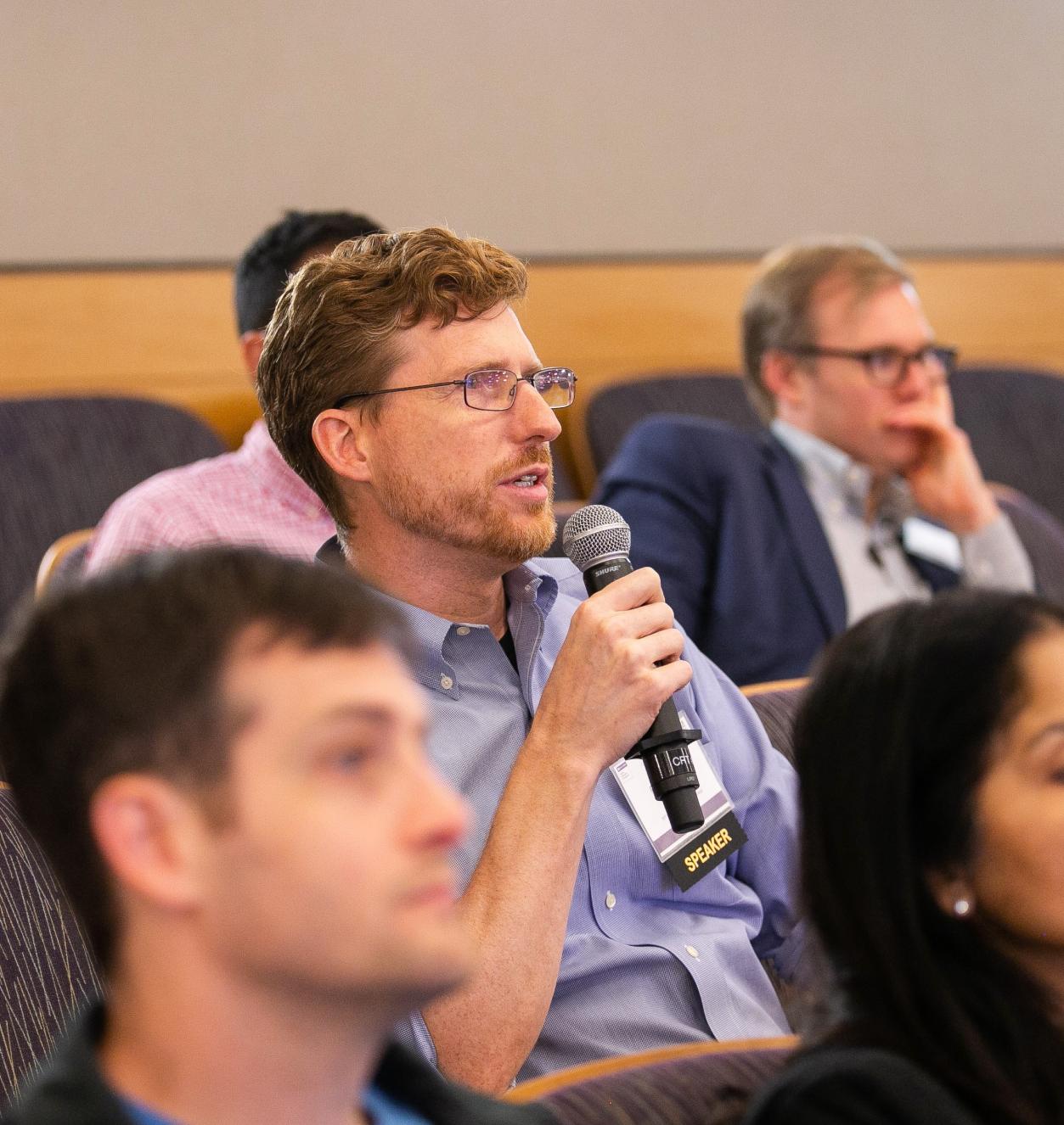 Robert Seamans sitting among the crowd in a lecture hall holding a microphone.