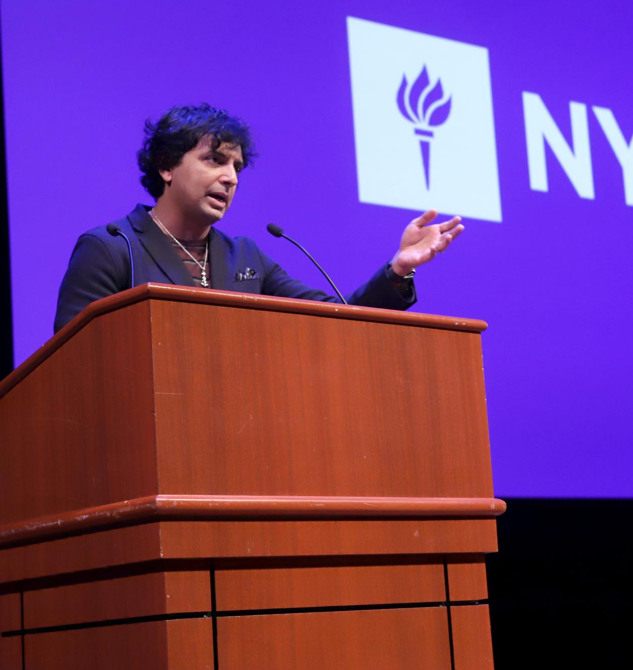 Filmmaker M. Night Shyamalan speaking at a podium in front of NYU logo background